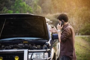 A man on a cell phone stands beside a broken car, making an emergency repair call for assistance. 